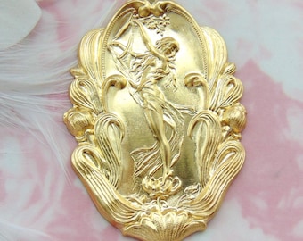 May include: A gold-toned metal pendant featuring a raised design of a woman holding a flower. The pendant is oval-shaped and has a detailed floral border.