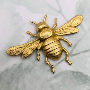 May include: A gold-colored bee brooch with detailed wings, legs, and body segments. The brooch is made of metal and has a textured surface. The bee is a classic insect design, perfect for adding a touch of nature-inspired flair to any outfit.