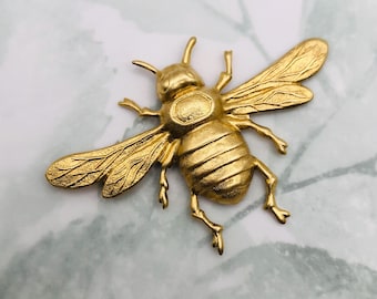 May include: A gold-colored bee brooch with detailed wings, legs, and body segments. The brooch is made of metal and has a textured surface. The bee is a classic insect design, perfect for adding a touch of nature-inspired flair to any outfit.