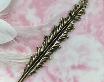 May include: A bronze-toned metal leaf-shaped embellishment with detailed design. The decorative piece has a textured surface and a pointed tip. The leaf is set against a soft pink and white background, suggesting a craft or jewelry component.