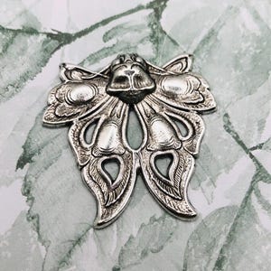 Puede incluir: Adorno de metal plateado con forma de mariposa. El diseño detallado presenta alas estilizadas y un cuerpo central con intrincados patrones florales y de hojas. La mariposa es un elemento decorativo.