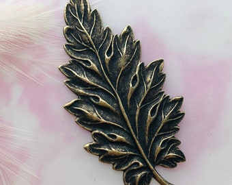 May include: A detailed, antique gold-toned metal leaf charm with a textured surface. The leaf has a realistic design with intricate veins and a pointed tip.
