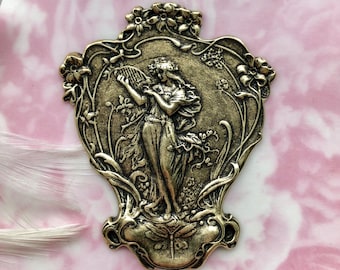 May include: A gold-toned metal pendant featuring a woman playing a lyre in a floral design. The pendant is in an ornate, art nouveau style.