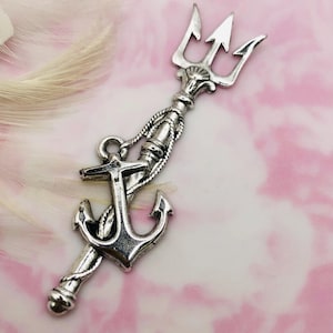 Puede incluir: Encanto de plata con un diseño de tridente y ancla. El tridente está en la parte superior del encanto y el ancla en la parte inferior. El encanto está sobre un fondo rosa y blanco.