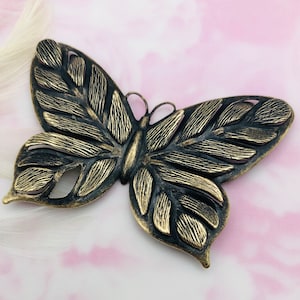 May include: A gold-toned metal butterfly charm with a detailed, textured design. The butterfly has a cutout design and is perfect for adding a touch of whimsy to jewelry or crafts.