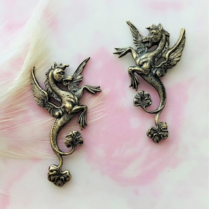 May include: Two antique bronze-colored seahorse-pegasus hybrid charms. Each charm features a winged horse with a seahorse tail, standing on its hind legs. The charms have intricate details and are set against a pink and white background.