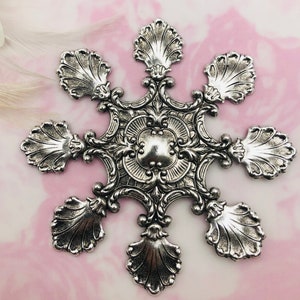 May include: A silver snowflake ornament with a detailed design featuring a central circular motif and six scalloped edges.