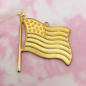 May include: A gold-colored metal American flag charm with a decorative finial on the top of the pole. The flag is waving in the wind.