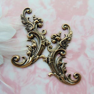 Op de afbeelding: Twee antieke, goudkleurige, decoratieve metalen versieringen met een scroll-ontwerp en een vogelmotief. De versieringen zijn sierlijk en gedetailleerd, met een vintage esthetiek.
