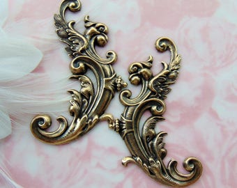 May include: Two antique gold-toned metal decorative embellishments with a scroll design and a bird motif. The embellishments are ornate and detailed, with a vintage aesthetic.