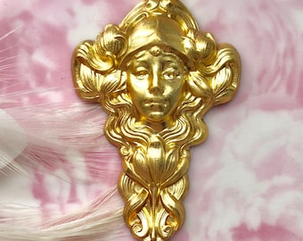 May include: A gold-colored, Art Nouveau-style decorative element featuring a woman's face with flowing hair and a helmet. The ornate design includes floral and scroll details, creating a vintage aesthetic. The piece is likely made of metal and has a polished finish.