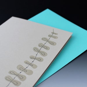 May include: A blank card with a light brown background and a simple design of a string of leaves in gray. The card is folded in half and is sitting on top of a turquoise envelope.