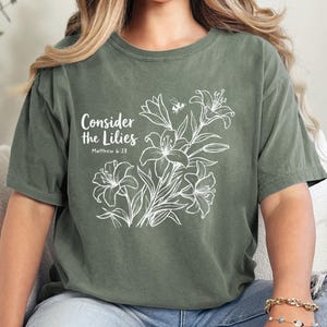 May include: Moss green t-shirt with the text "Consider the Lilies" and a floral illustration. The design includes white lilies and a small bee. The shirt is made of a soft material and is a comfortable fit.