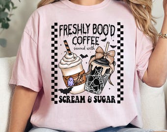 May include: A light pink t-shirt with a Halloween-themed graphic. The graphic features two coffee drinks with whipped cream, bats, and a spiderweb design. Text reads "FRESHLY BOO'D COFFEE served with SCREAM & SUGAR."