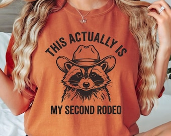 May include: Burnt orange t-shirt with a raccoon wearing a cowboy hat graphic. The text reads "THIS ACTUALLY IS MY SECOND RODEO" in black. The shirt is a comfortable, casual style.