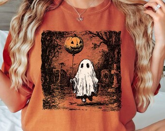 May include: Orange-colored sweatshirt featuring a Halloween-themed graphic. The graphic depicts a ghost holding a pumpkin balloon in a graveyard setting. The design is in black and white, with the ghost and balloon in the foreground.