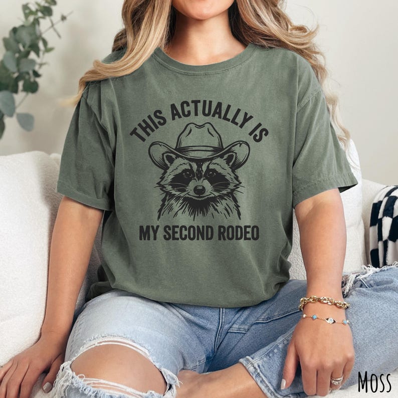 Comfort Colors moss green t-shirt with “This Actually Is My Second Rodeo” text and a raccoon wearing a cowboy hat graphic.