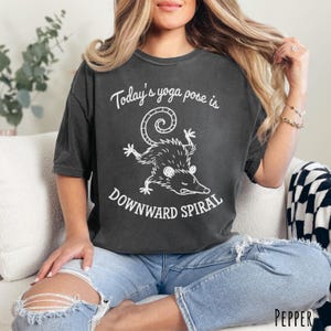 Pepper Comfort Colors unisex T-shirt with “Today’s yoga pose is Downward Spiral” cartoon possum graphic in black line art.