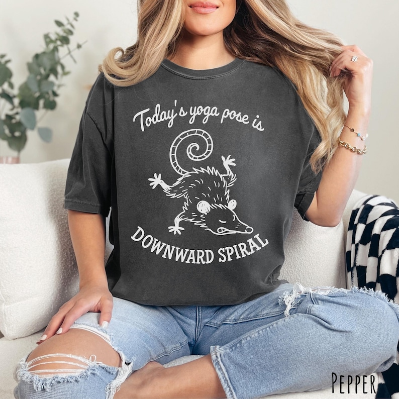 Pepper Comfort Colors unisex T-shirt with “Today’s yoga pose is Downward Spiral” cartoon possum graphic in black line art.
