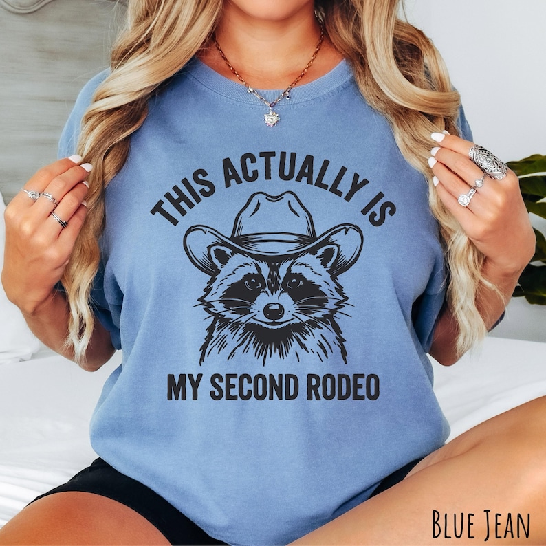 Comfort Colors blue jean t-shirt with “This Actually Is My Second Rodeo” text and a raccoon wearing a cowboy hat graphic.