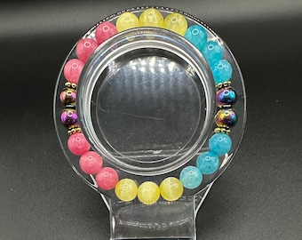 May include: A bracelet with pink, yellow, and blue beads, with iridescent silver beads interspersed. The bracelet is displayed on a clear plastic stand. A sticker with the text "PANSEXUAL PRIDE" and the pansexual flag colors is shown below the bracelet.