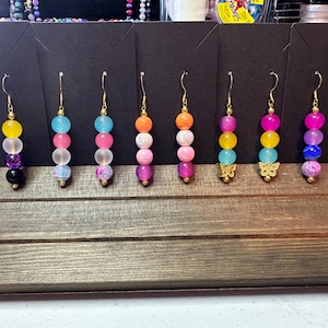 May include: A display of eight pairs of dangle earrings with colorful beads. Each pair features a different color combination of beads, including yellow, pink, blue, purple, and orange. Some of the earrings have a butterfly charm at the bottom.