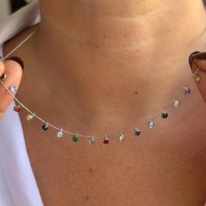 Könnte beinhalten: Eine silberne Halskette mit einer filigranen Kette, verziert mit kleinen, bunten Edelsteinen. Die Edelsteine sind in verschiedenen Farben gehalten, darunter Rot, Gelb, Grün und Blau. Die Halskette wird hochgehalten, um ihr Design und die leuchtenden Farben der Steine zu zeigen.