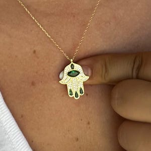 May include: A gold-colored necklace featuring a Hamsa hand pendant. The pendant is adorned with small, clear stones and green gemstones. The necklace is worn against a white shirt.