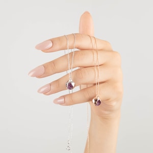 May include: Two delicate silver and gold necklaces with a small, round pendant featuring a purple gemstone. The necklaces are held in a hand with light pink nail polish.