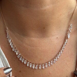 May include: A delicate gold-toned necklace featuring a series of clear, rectangular-cut crystals. The crystals are evenly spaced along the necklace's curve, creating a sparkling, elegant design. The necklace is worn around the neck.