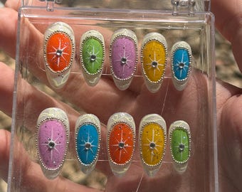 Puede incluir: Un juego de diez uñas postizas ovaladas en una caja de plástico transparente. Cada uña presenta una base blanca con un diseño colorido: naranja, verde, rosa, amarillo y azul. El diseño incluye una estrella y un borde de cuentas plateadas.