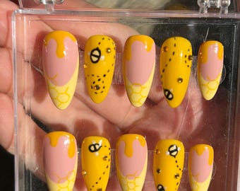 May include: A set of ten artificial nails in a clear plastic case. The nails are yellow with pink and honey-colored designs. Some nails feature black bee graphics and small rhinestones. The nails are oval-shaped.