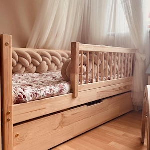 Könnte beinhalten: Ein hellfarbiger Holzbettenrahmen mit einer Matratze mit floralem Muster. Das Kopfteil hat ein geflochtenes Design, und das Bett hat eine ausziehbare Schublade. Ein transparenter Vorhang hängt im Hintergrund und schafft eine weiche, einladende Atmosphäre.