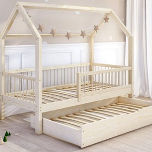 Könnte beinhalten: Ein weißes, hausförmiges Bettgestell aus Holz mit einer herausziehbaren Schublade. Das Bettgestell hat ein Lattenkopfteil und ein Fußteil sowie eine dekorative Sternen-Girlande oben.