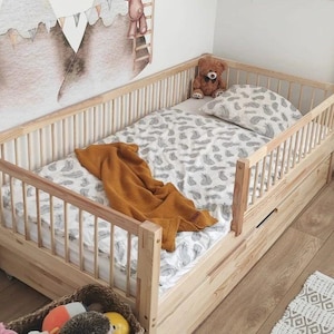 Könnte beinhalten: Ein helles Holzbettchen mit einem Feder-Muster-Bettlaken und Kissen. Eine senffarbene Decke ist über das Bett drapiert. Ein brauner Teddybär sitzt auf dem Kissen. Eine Schublade ist in den Bettrahmen eingebaut.