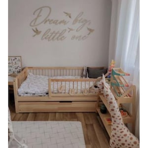 Könnte beinhalten: Ein helles Holzbett für Kleinkinder mit einer Schublade, einer gemusterten Bettdecke und einem Sicherheitsgeländer. Ein Giraffen-Spielzeug und ein Holzregal mit Spielzeug befinden sich neben dem Bett. An der Wand steht der Text "Dream big, little one."