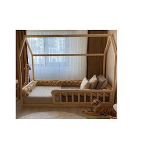 Peut inclure: Un lit en bois en forme de maison avec un profil bas. Le lit est doté d'un pare-chocs tressé et est accessoirisé avec des oreillers et une girafe et un tigre en peluche. Le cadre de lit est conçu pour une chambre d'enfant.
