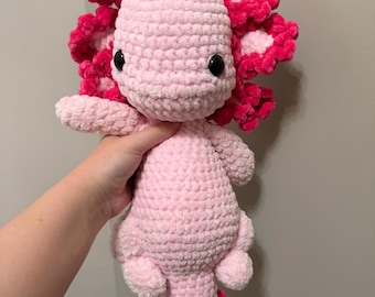May include: A plush axolotl toy made of light pink yarn with a textured, crocheted finish. It features large black plastic eyes, prominent dark pink external gills on both sides of the head, and a matching dark pink tail fin at the bottom.