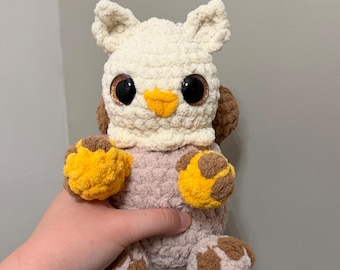 May include: A handmade crochet plush toy featuring a cream-colored head with pointed ears, large black glossy eyes, and a small yellow beak. The body is light brown with textured paws in yellow and brown, and a fluffy brown tail visible behind.