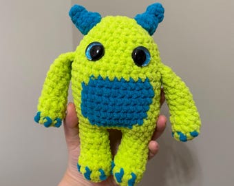 May include: A handmade crochet monster plush toy featuring a bright lime green body with contrasting blue accents on the horns, belly patch, hands, and feet. The creature has two large, round, black plastic safety eyes with blue glittery rims.