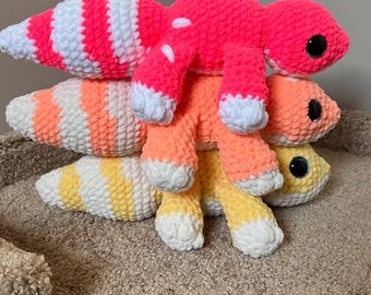 May include: Stack of three crocheted gecko plushies in vibrant colors. The top gecko is bright pink with white spots and stripes. The middle gecko is orange with white accents, and the bottom gecko is yellow with white stripes. Each has large, black, round eyes.