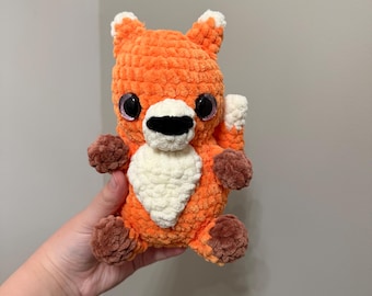 May include: A small, handmade crochet fox plush toy made from orange, white, and brown yarn. The fox features large, dark, glossy safety eyes, a black stitched nose, and a white chest patch. It has brown paws and a white-tipped tail.