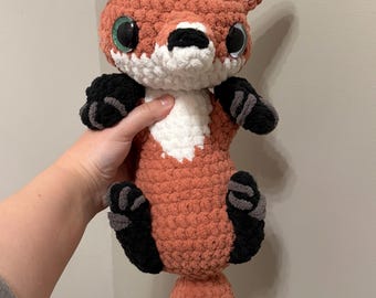 May include: A handmade crochet fox plush toy featuring a rust-orange body, a white chest patch, and white-tipped ears. It has large, round, dark eyes with green accents, a black nose, and black paws with grey detailing on a long, thick tail.