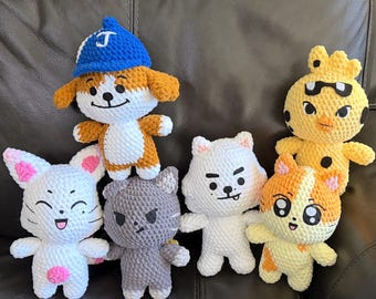 May include: A collection of six handmade crochet plush toys featuring various characters. The group includes a brown and white dog wearing a blue baseball cap with a white letter J, a white cat, a gray cat, a white creature, a yellow bird, and a calico cat.