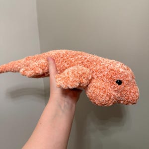 May include: A plush toy shaped like a lizard or salamander, crafted from fuzzy, textured yarn in a variegated blend of orange and white. The creature features a long, slender tail, four short legs, and a single small, round, black plastic eye.