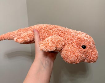 May include: A plush toy shaped like a lizard or salamander, crafted from fuzzy, textured yarn in a variegated blend of orange and white. The creature features a long, slender tail, four short legs, and a single small, round, black plastic eye.