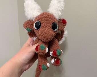 May include: A brown crocheted plush creature with large black eyes, white fuzzy ears, and a long tail. The body features small patches of red and green yarn accents on the feet and sides, creating a textured appearance with soft yarn.