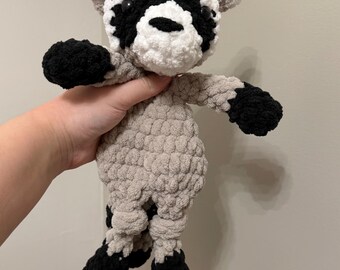 May include: A handmade crochet raccoon plush toy crafted from soft, chunky yarn. The raccoon features a light beige body, black paws, and a distinctive white and black facial mask with dark, round plastic eyes and a small black nose.