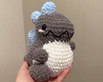 May include: A handmade crochet dinosaur plushie in shades of gray, white, and light blue. The dinosaur has a textured body, a white belly, and blue spikes along its back. It features black eyes and a small pink detail.