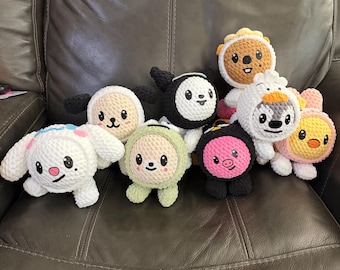 May include: A collection of eight small, round, crocheted plush toys featuring various animal-inspired designs. Each figure has a soft, textured yarn surface, embroidered facial features, and distinct color schemes including white, black, pink, green, and orange.